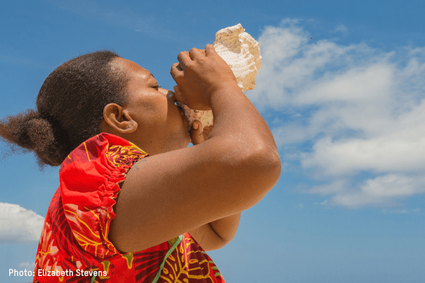 A woman blows a conch shell toward the sky