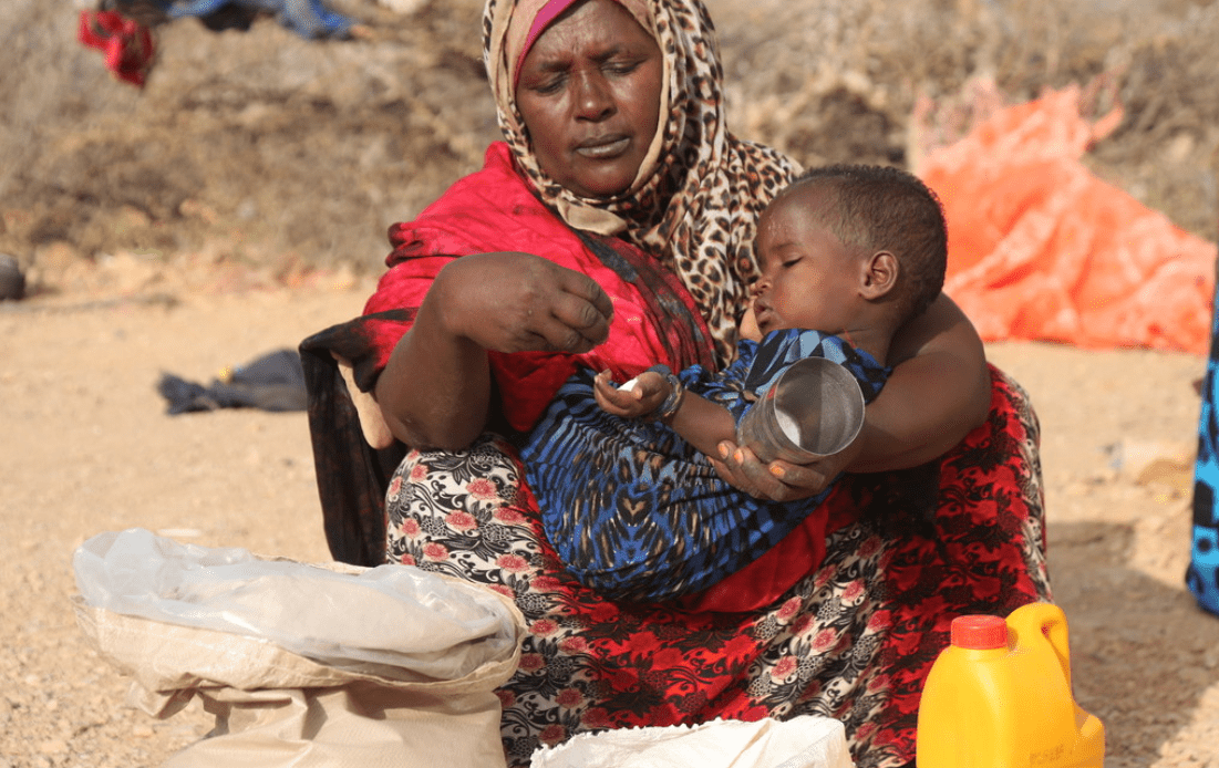 A woman holds her young child, feeding from from a large bag of grain.